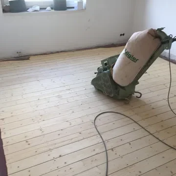 Holzboden mit einer Schleifmaschine in einem lichtdurchfluteten Raum.