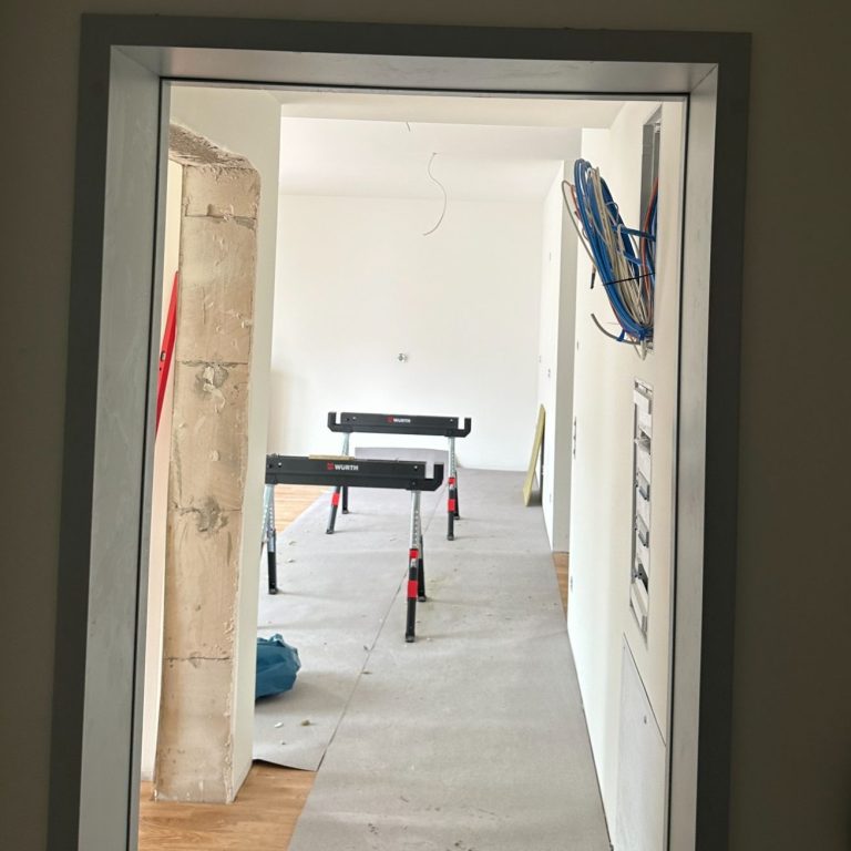 Blick durch eine Tür auf einen leeren Raum mit Baugeräten und Bodenbelag.